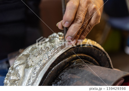 Close up of artisan hand engraving detailed pattern on metal surface using carving tool traditional craft skill craftsmanship jewelry making workshop process 139429398