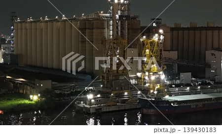 A global maritime grain terminal with massive concrete silos, illuminated yellow cranes, and the bulk carrier ship docked for loading under floodlights 139430183