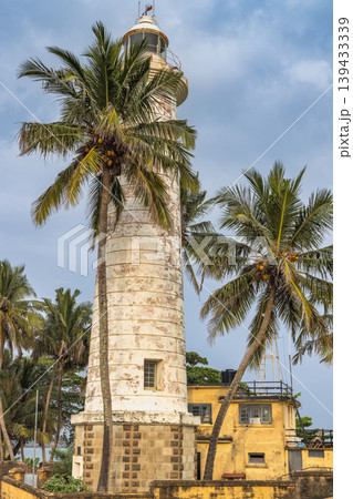 Historic white lighthouse tower stands tall amidst tropical palm trees under a bright blue sky, coastal architecture. Galle, the historic port city on the southwest coast of Sri Lanka. Historic white lighthouse tower stands tall amidst tropical palm trees under a bright blue sky, coastal architecture. Galle, the historic port city on the southwest coast of Sri Lanka. 139433339