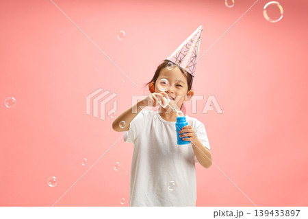 Cheerful Female Kid Around 5-6 Years Old Happy Blowing Soap Bubbles Wearing in Paper Party Hat and White T Shirt Standing on Pink Isolated Background Cheerful Female Kid Around 5-6 Years Old Happy Blowing Soap Bubbles Wearing in Paper Party Hat and White T Shirt Standing on Pink Isolated Background 139433897