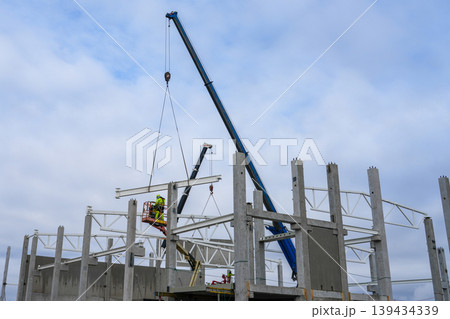 Crane lifting steel beam while worker on lift guides placement on concrete frame at build site Crane lifting steel beam while worker on lift guides placement on concrete frame at build site 139434339