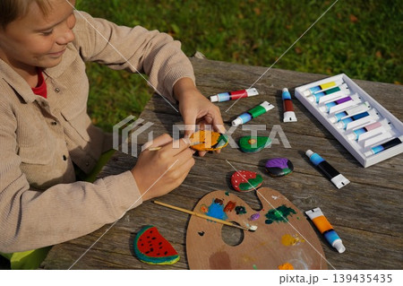 A child decorating flat pebbles with acrylic paint, depicting fresh produce like apples, carrots, and tomatoes. A child decorating flat pebbles with acrylic paint, depicting fresh produce like apples, carrots, and tomatoes. 139435435