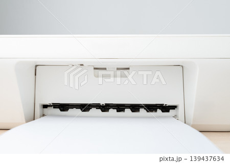 Close-up view of a white printer feeding blank paper into the output tray, suggesting office printing, document production, minimal workspace and modern office equipment for home or business use. 139437634