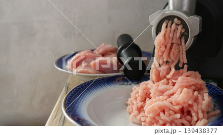 A black electric meat grinder and two pink plates with poultry fillet and ready-made minced meat, ground on a household appliance, the meat grinder in operation on an empty background of a light wall A black electric meat grinder and two pink plates with poultry fillet and ready-made minced meat, ground on a household appliance, the meat grinder in operation on an empty background of a light wall 139438377