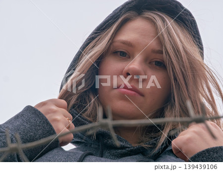 Young woman with an intense gaze in a dark hoodie behind a barbed wire fence on an overcast day Young woman with an intense gaze in a dark hoodie behind a barbed wire fence on an overcast day 139439106