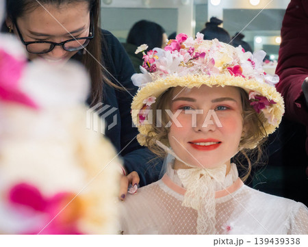 Smiling woman in elaborate floral hat being prepared by makeup artist in a brightly lit dressing room Smiling woman in elaborate floral hat being prepared by makeup artist in a brightly lit dressing room 139439338