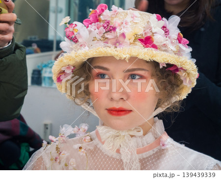Young woman wearing elaborate floral straw hat and white lace top with artistic makeup prepares for a performance indoors Young woman wearing elaborate floral straw hat and white lace top with artistic makeup prepares for a performance indoors 139439339
