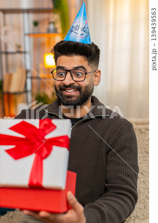 Indian man on sofa opens birthday gift with smile, showing joy and surprise in cheerful celebration Indian man on sofa opens birthday gift with smile, showing joy and surprise in cheerful celebration 139439563