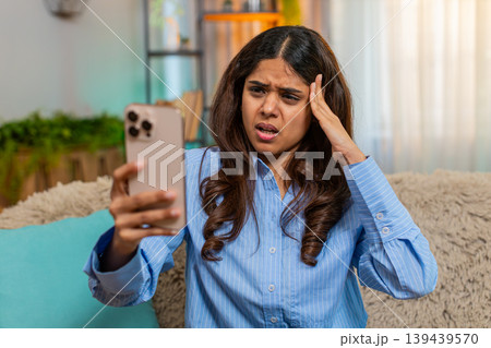 Indian woman looking down with sad sigh while reading bad news on smartphone on home sofa indoors 139439570