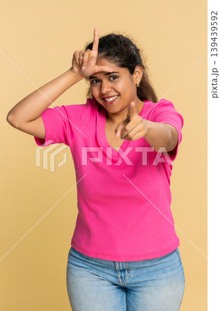 Indian woman making L loser sign on forehead, smiling ironically, humorously teasing someone Indian woman making L loser sign on forehead, smiling ironically, humorously teasing someone 139439592