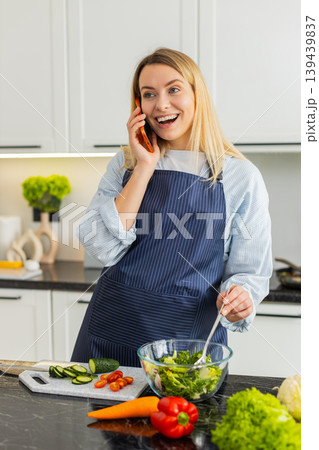 Young woman talking on orange smartphone while mixing salad in kitchen sharing healthy keto recipe 139439837