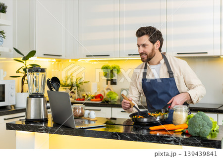 Middle-aged man cooking vegetables on electric stove while watching laptop recipe for healthy diet 139439841