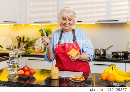 Senior old woman dancing with headphones while making peanut butter toast in home kitchen for diet Senior old woman dancing with headphones while making peanut butter toast in home kitchen for diet 139439877