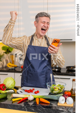 Middle-aged man celebrating victory on smartphone in kitchen during healthy meal preparation at home Middle-aged man celebrating victory on smartphone in kitchen during healthy meal preparation at home 139439894