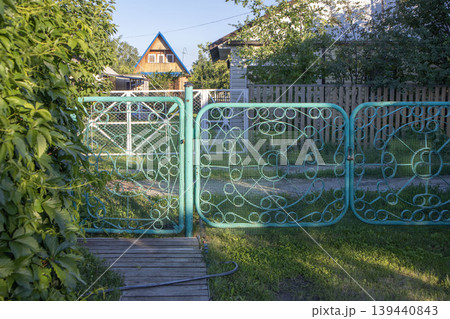 Decorative turquoise metal gate with intricate patterns leading to a garden path, surrounded by greenery and a wooden walkway in a residential area Decorative turquoise metal gate with intricate patterns leading to a garden path, surrounded by greenery and a wooden walkway in a residential area 139440843