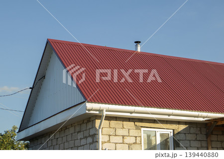 Red metal roof with vertical ribbing on a stone house under clear blue sky, chimney visible on the roofline, showcasing modern architectural design elements 139440880