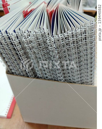 Stack of spiral-bound notebooks with colorful covers arranged neatly in a white storage box on a wooden surface in an office or study environment Stack of spiral-bound notebooks with colorful covers arranged neatly in a white storage box on a wooden surface in an office or study environment 139440882
