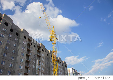 Yellow construction crane towering over unfinished apartment building against blue sky Yellow construction crane towering over unfinished apartment building against blue sky 139441482
