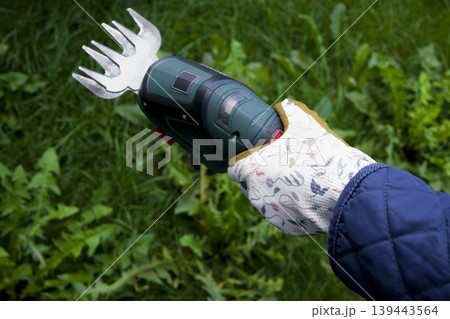 Woman's hand holds a green electric grass shears among grass, weeds and small wildflowers in garden Woman's hand holds a green electric grass shears among grass, weeds and small wildflowers in garden 139443564