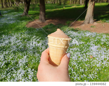 A woman's hand holds a waffle cup with chocolate ice cream against the background of a bright spring meadow 139444082