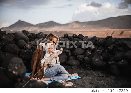 Mother enjoying winter vacations playing with his infant baby boy son on black sandy volcanic beach of Janubio on Lanzarote island, Spain on windy overcast day. Family travel vacations concept Mother enjoying winter vacations playing with his infant baby boy son on black sandy volcanic beach of Janubio on Lanzarote island, Spain on windy overcast day. Family travel vacations concept 139447239