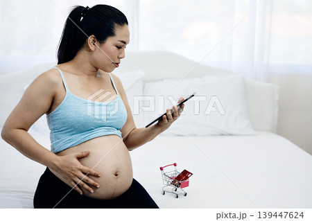 Pregnant woman using smartphone for online shopping while touching baby bump at home, digital maternity lifestyle and ecommerce payment concept for modern motherhood and prenatal preparation 139447624