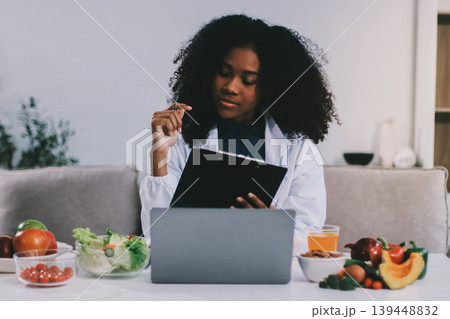 Young nutritionist showing bell peppers during an online video call consulting about healthy eating, sitting at home at a table with vegetables, laptop and measuring tape Young nutritionist showing bell peppers during an online video call consulting about healthy eating, sitting at home at a table with vegetables, laptop and measuring tape 139448832