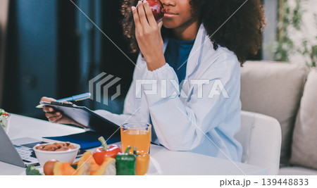 Young nutritionist showing bell peppers during an online video call consulting about healthy eating, sitting at home at a table with vegetables, laptop and measuring tape 139448833