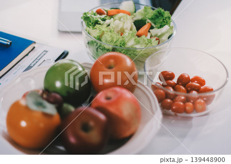 Young nutritionist showing bell peppers during an online video call consulting about healthy eating, sitting at home at a table with vegetables, laptop and measuring tape 139448900