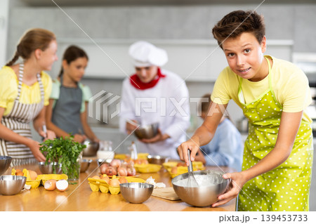 During cooking classes, teen boy carefully mixes ingredients in bowl with whisk During cooking classes, teen boy carefully mixes ingredients in bowl with whisk 139453733