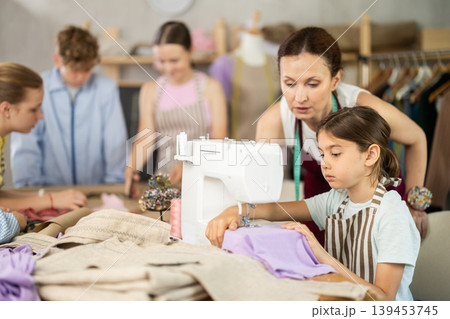 Teenage girl tries to sew on sewing machine. Teacher helps. Children watch and learn 139453745