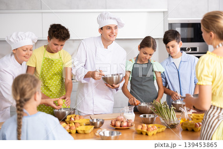 During cooking lesson, male cook tells children about culinary rules, woman assistant helps child During cooking lesson, male cook tells children about culinary rules, woman assistant helps child 139453865