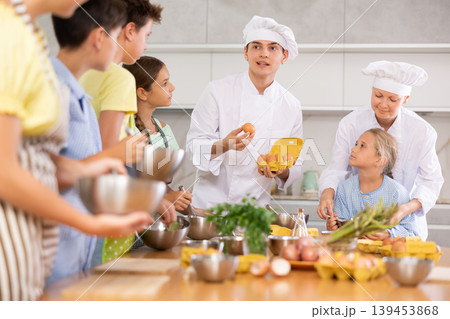 During cooking lesson, male cook tells children about culinary rules, woman assistant helps child During cooking lesson, male cook tells children about culinary rules, woman assistant helps child 139453868