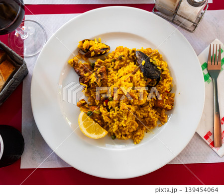 Paella made of rice, chicken and different seafood served in white plate with table appointments 139454064