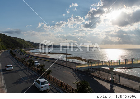 海岸線沿いの道路に架かる横断歩道橋と雲の隙間から四方に光が差す海上 139455666