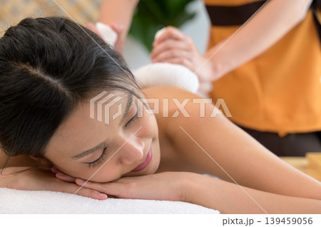 Asian young woman with eyes closed enjoying professional back massage with Thai herbal compress on massage table at spa salon Asian young woman with eyes closed enjoying professional back massage with Thai herbal compress on massage table at spa salon 139459056