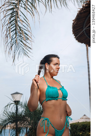 Attractive woman touching braided hair with radiant bronzed healthy skin beside tropical palms outdoors. Attractive female showing smooth glowing body texture during summer resort vacation and seaside Attractive woman touching braided hair with radiant bronzed healthy skin beside tropical palms outdoors. Attractive female showing smooth glowing body texture during summer resort vacation and seaside 139468427