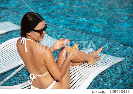 Young woman applying skincare lotion on smooth glowing legs beside tropical swimming pool outdoors. Beautiful female showing radiant healthy skin texture during luxury summer vacation near seaside 139468458