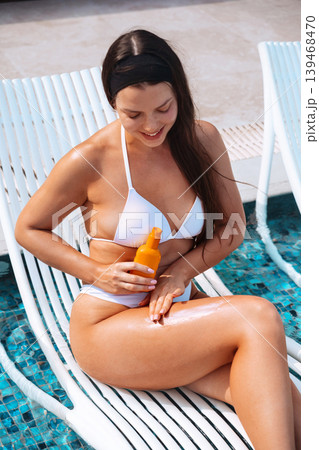 Young woman using sunscreen lotion with glowing healthy complexion beside tropical swimming pool outdoors. Beautiful female applying sun cream and holding skincare bottle while showing smooth radiant 139468470