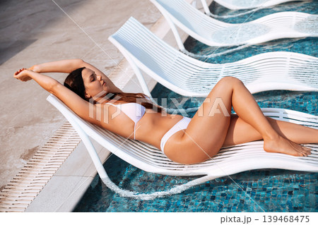 Beautiful woman relaxing on floating pool chair with glowing bronzed summer skin outdoors. Beautiful female posing in white bikini showing smooth radiant healthy body texture beside luxury swimming Beautiful woman relaxing on floating pool chair with glowing bronzed summer skin outdoors. Beautiful female posing in white bikini showing smooth radiant healthy body texture beside luxury swimming 139468475