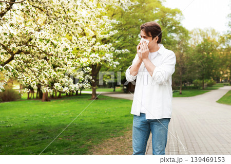 Man in a park blowing his nose with a tissue due to seasonal allergies. Concept of hay fever, pollen sensitivity, and rhinitis symptoms during the spring blooming season. 139469153