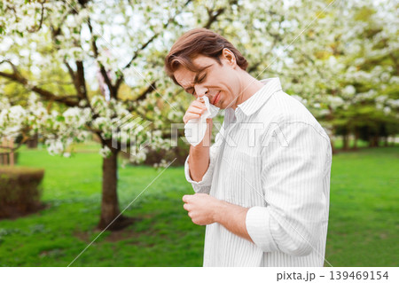 Man rubbing his itchy eyes with a tissue while standing in a park with blooming trees. Concept of severe seasonal allergy, pollen sensitivity, and hay fever during spring. 139469154