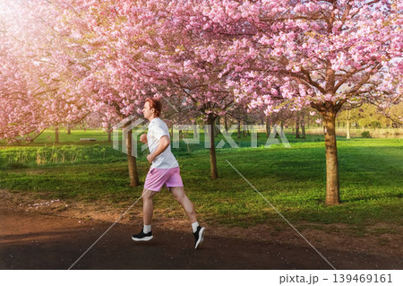 Side view of a man jogging through a park under a canopy of blooming pink cherry blossoms. A vibrant spring morning scene promoting fitness, health, and outdoor activity. 139469161