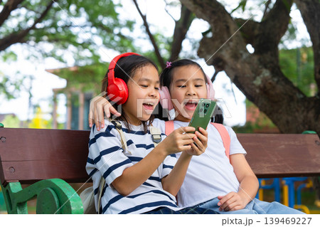 Two Asian student girls wearing headphones and using a smartphone together while sitting on a park bench, Back to school and digital lifestyle concept. Two Asian student girls wearing headphones and using a smartphone together while sitting on a park bench, Back to school and digital lifestyle concept. 139469227