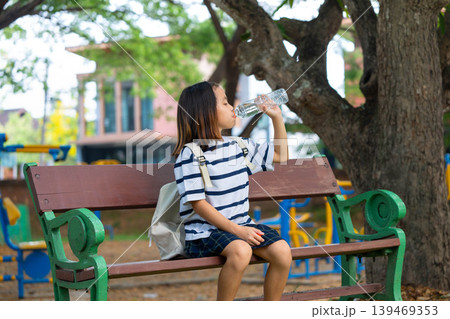 Cute Asian student girl sitting on a park bench and drinking water from a plastic bottle, Back to school and healthy lifestyle concept. Cute Asian student girl sitting on a park bench and drinking water from a plastic bottle, Back to school and healthy lifestyle concept. 139469353