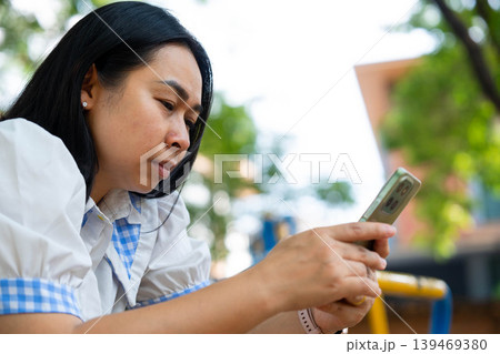 Asian mother using a smartphone while sitting on a bench and waiting for her children at the school park, Back to school and lifestyle concept. 139469380