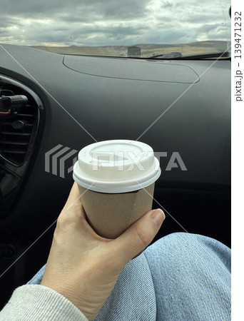 Car ride with hand holding coffee cup inside vehicle against scenic road background Car ride with hand holding coffee cup inside vehicle against scenic road background 139471232