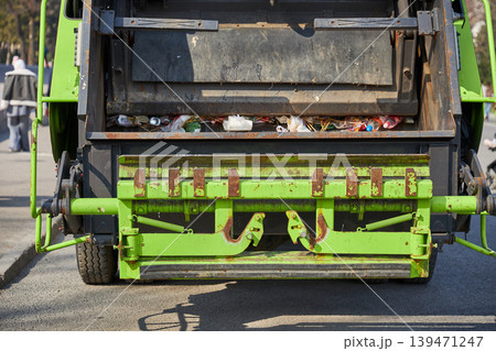 Rear view of green garbage truck collecting waste on city street. Industrial vehicle for trash disposal and city sanitation service. Management of urban municipal solid waste recycling. Rear view of green garbage truck collecting waste on city street. Industrial vehicle for trash disposal and city sanitation service. Management of urban municipal solid waste recycling. 139471247