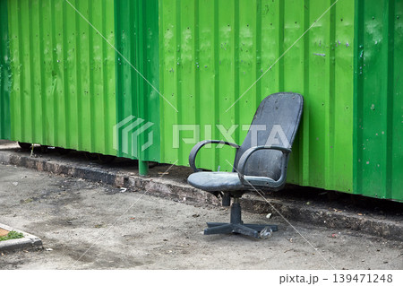 Old black office chair abandoned on street near green metal wall. Concept of unemployment, discarded furniture and office closure. Urban decay and lonely object in city environment. Old black office chair abandoned on street near green metal wall. Concept of unemployment, discarded furniture and office closure. Urban decay and lonely object in city environment. 139471248