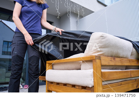 Woman places black protective cover over wooden patio sofa with beige cushions Woman places black protective cover over wooden patio sofa with beige cushions 139471750